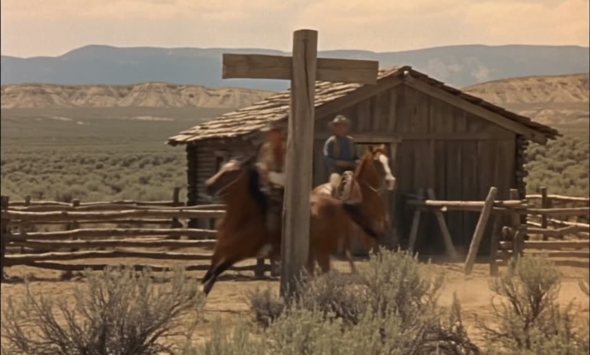 A rider at full gallop past a frontier relay cabin and corral